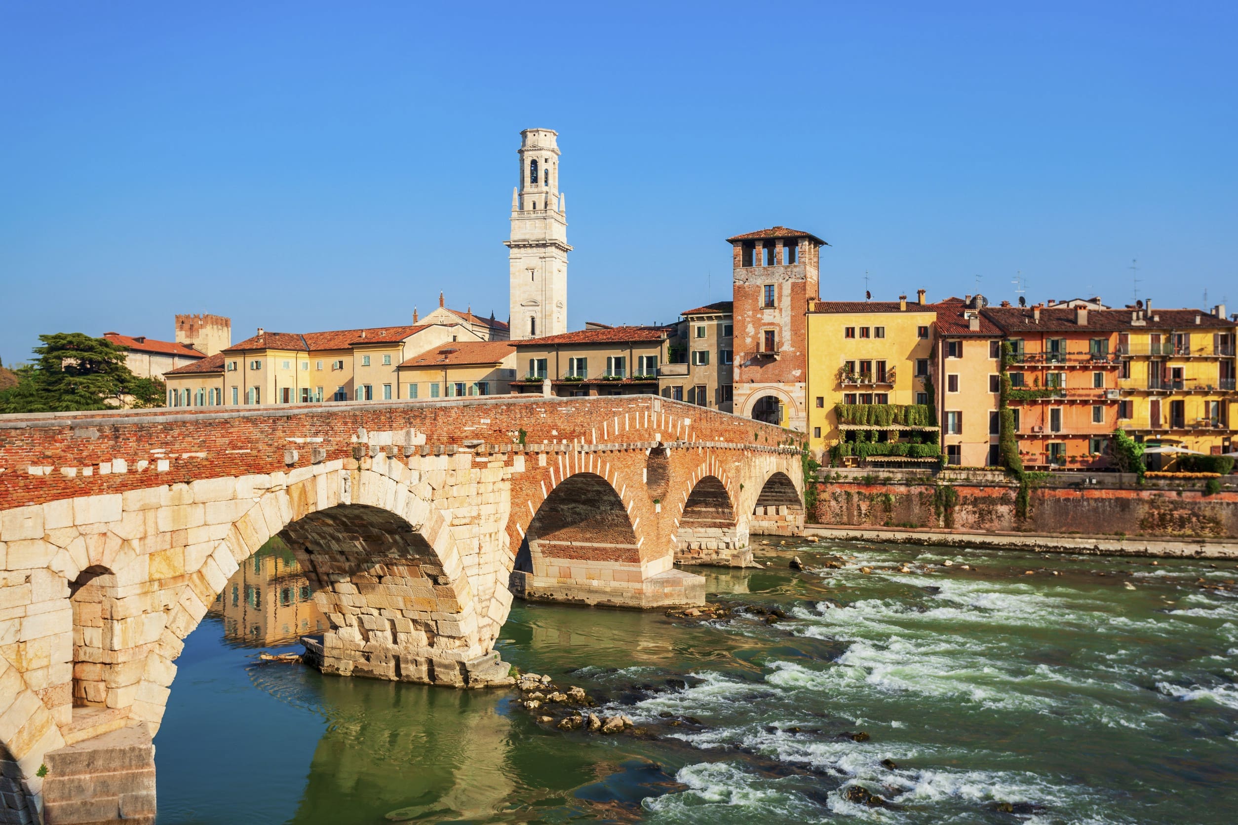 「義大利維洛納(Verona)的著名景點——佩雅托橋(Ponte Pietra)。這是一座橫跨阿迪傑河(Adige River)的古老羅馬拱橋,由紅磚和石塊砌成。背景是維洛納老城的彩色房屋,以及一座醒目的白色高塔(可能是蘭貝爾提塔或聖彼得城堡的塔樓),河水在橋下湍急流動。」