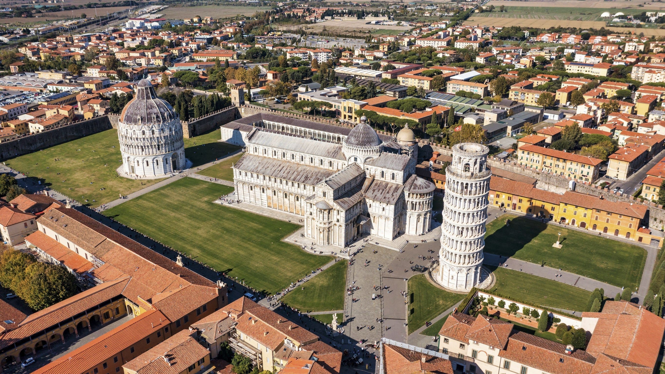 義大利比薩奇蹟廣場(Piazza dei Miracoli)的俯瞰圖。畫面中心是廣闊的綠色草坪,上面坐落著三座標誌性的白色大理石建築:最右側是明顯傾斜的比薩斜塔(鐘樓);中間是宏偉的比薩主教堂(Duomo di Pisa);最左側是圓頂的洗禮堂(Baptistery)。整個廣場被周圍密集的橘紅色屋頂老城區所環繞,清晰地展現了這處世界文化遺產與城市景觀的對比。