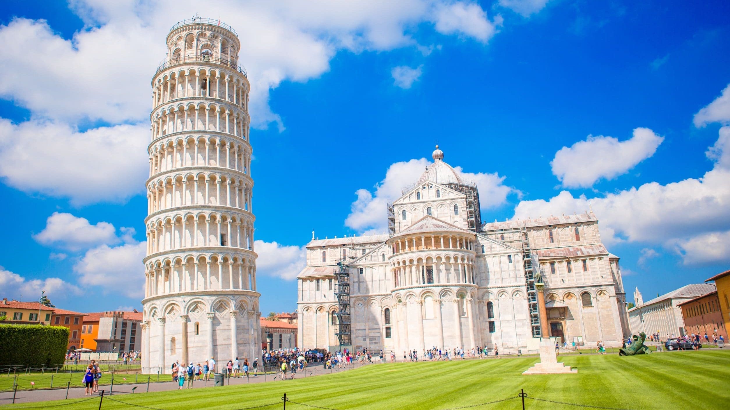 義大利比薩奇蹟廣場(Piazza dei Miracoli)的景觀。畫面左側是著名的比薩斜塔(Leaning Tower of Pisa),其傾斜的圓柱形身軀在藍天白雲下清晰可見。斜塔右側是比薩主教堂(Duomo di Pisa)的側面和圓頂。兩座建築均由白色大理石砌成,前方是修剪整齊的翠綠草坪,草坪上和周圍可見許多遊客。