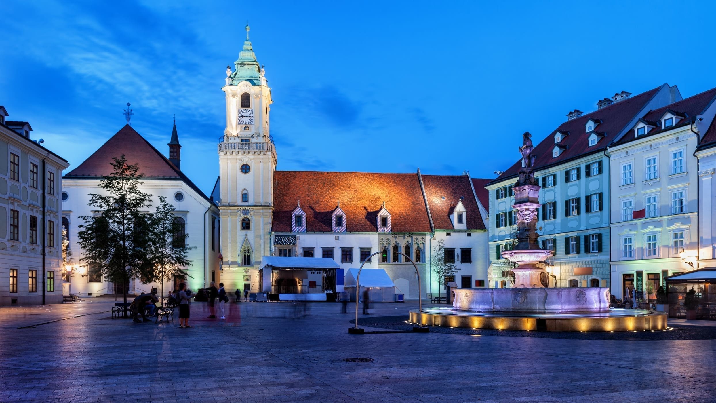 一張於黃昏或夜晚拍攝的斯洛伐克布拉提斯拉瓦 (Bratislava) 老城區主廣場 (Hlavné námestie) 照片。

廣場由鵝卵石鋪成，被周圍的歷史建築所環繞。中央是著名的羅蘭噴泉 (Roland Fountain)，噴泉被柔和的燈光點亮。在後方，矗立著帶有尖塔和時鐘的舊市政廳，其建築物有明顯的尖頂和紅瓦屋頂。旁邊還有其他傳統的歐式建築，它們的窗戶和立面也被點亮。天空呈現出深邃的藍色調，光線主要來自建築物和噴泉的人造光源。廣場上有幾個人影因長時間曝光而呈現模糊狀態。
